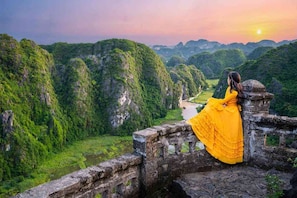 Una persona con un vestido amarillo sentada sobre un muro de piedra, con vistas a un valle exuberante con un río y montañas al fondo.