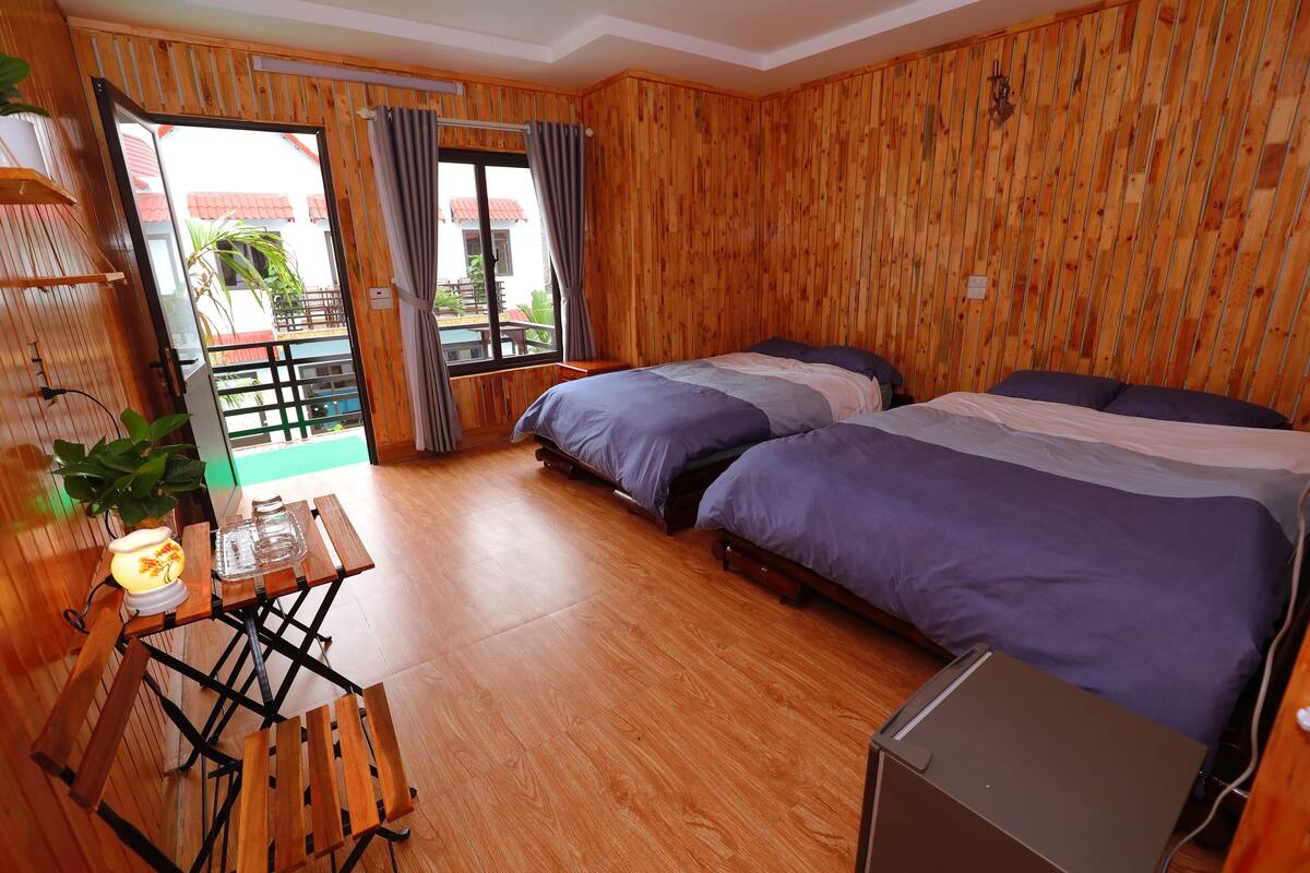 A room with wooden walls, two beds, a small table with a plant, and a view of a building outside.
