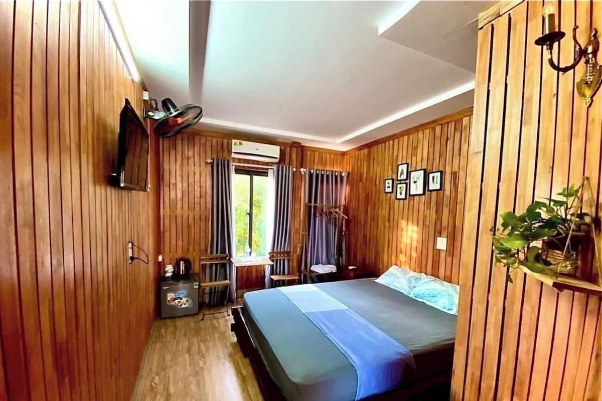 A room with wooden paneling, a bed with a blue bedspread, a small table with a chair, a wall-mounted TV, and a window with curtains.