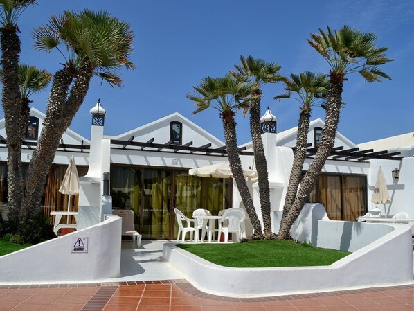 Un hôtel moderne aux murs extérieurs blancs, agrémenté de palmiers et doté d’un patio avec des chaises et des parasols blancs.