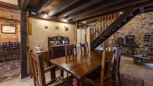 Uma área de jantar rústica com uma mesa e cadeiras de madeira, uma parede de pedra e uma escadaria que dá acesso a um andar superior.