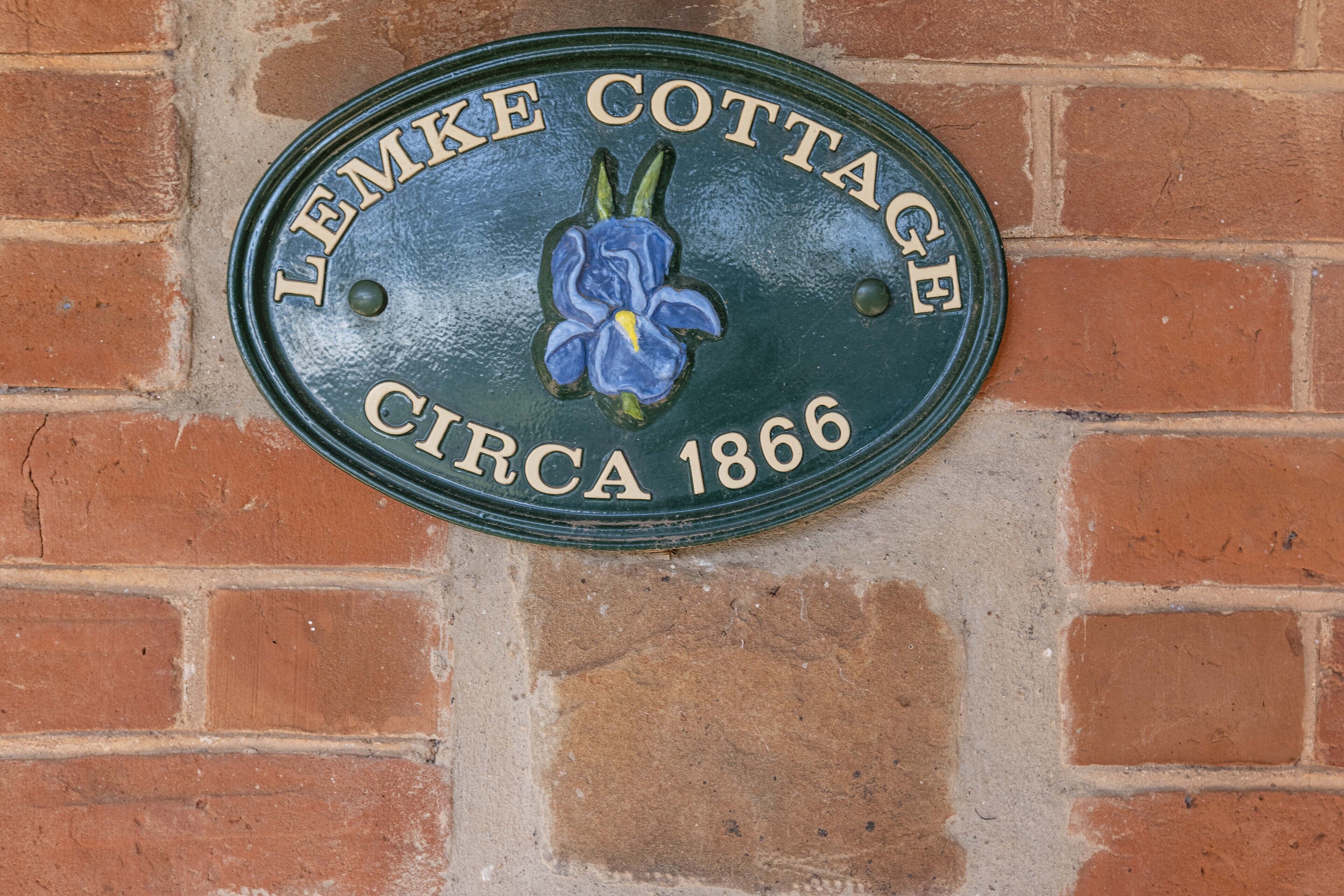 Een metalen plaquette met de inscriptie 'Lemke Cottage circa 1866' bevestigd aan een bakstenen muur.