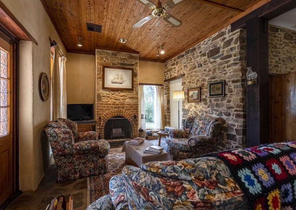 A cozy living room with a stone fireplace, two floral armchairs, a television, and a ceiling fan.