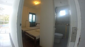 A room with a bed, a small table, a window with curtains, and a bathroom with a toilet and shower.
