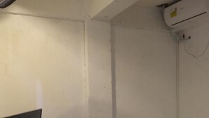 A small, dimly lit room with a single bed, a television on the left, and an air conditioning unit on the wall.