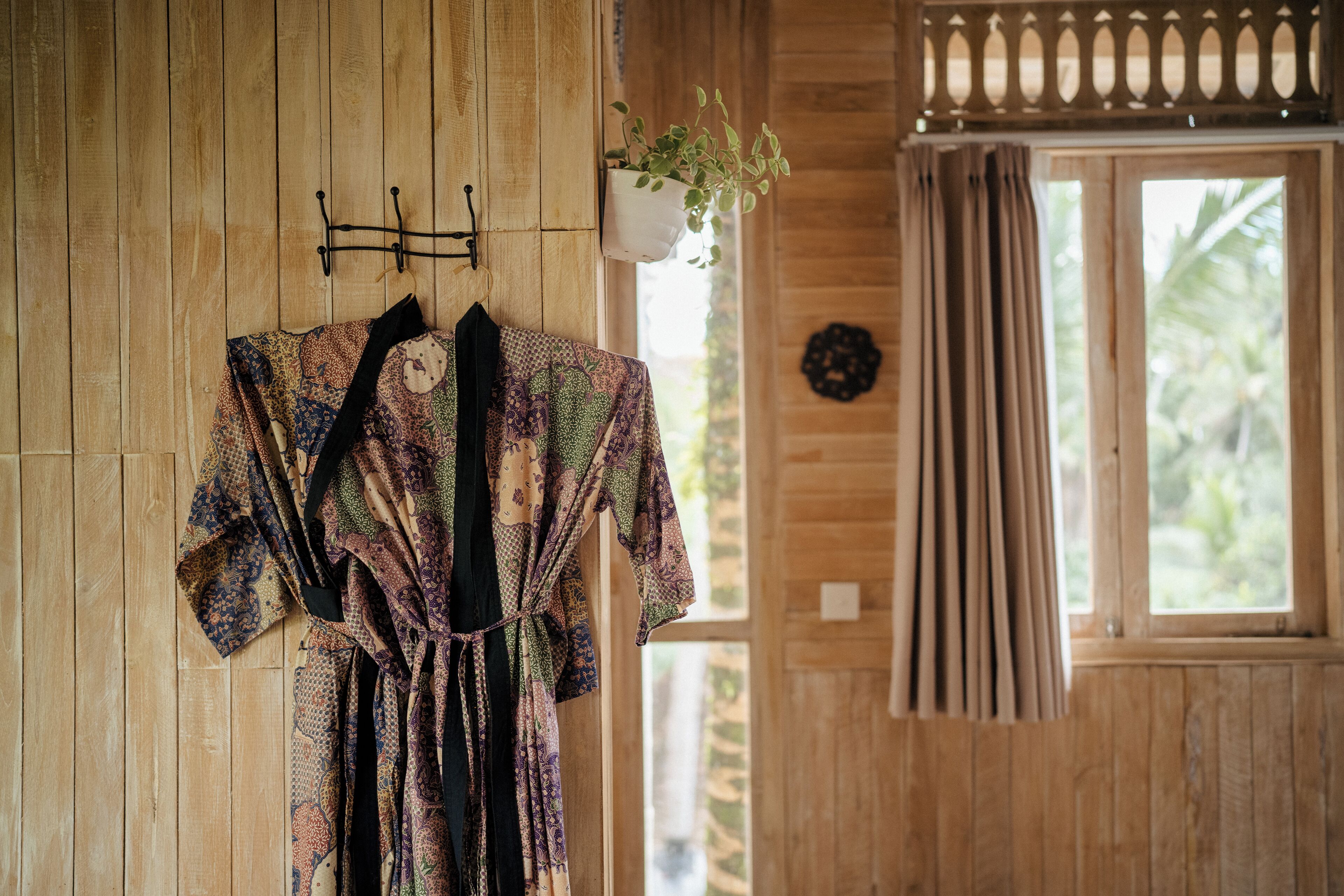 Une pièce en bois, avec une robe à motifs suspendue à un porte-vêtements fixé au mur, une plante en pot et une fenêtre avec des rideaux.