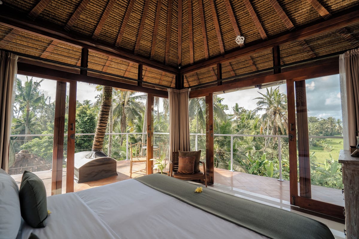 Quarto com cama, uma cadeira e vista para palmeiras.