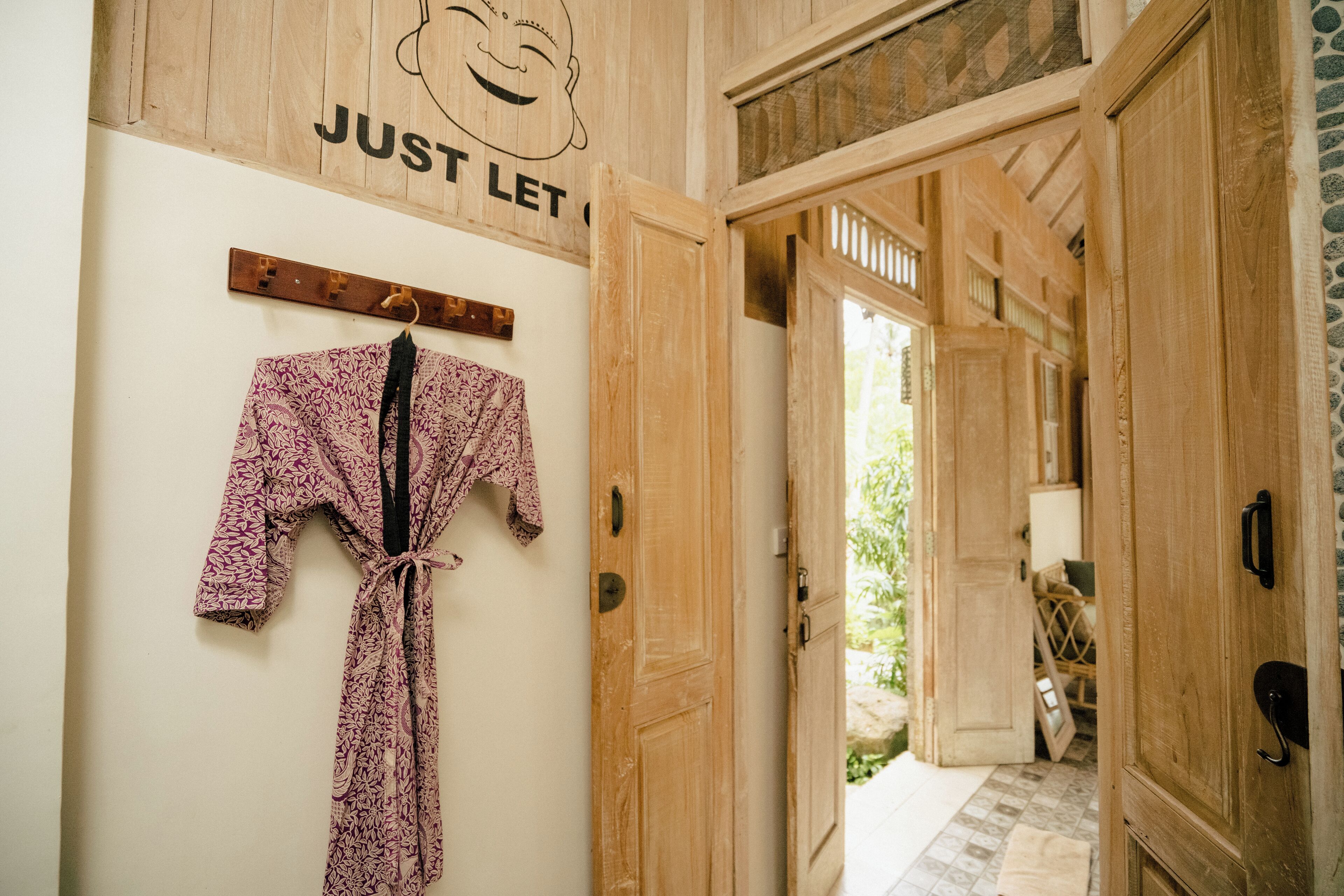A wooden door with a sign above it, a robe hanging on a rack, and a view of a garden through an open door.