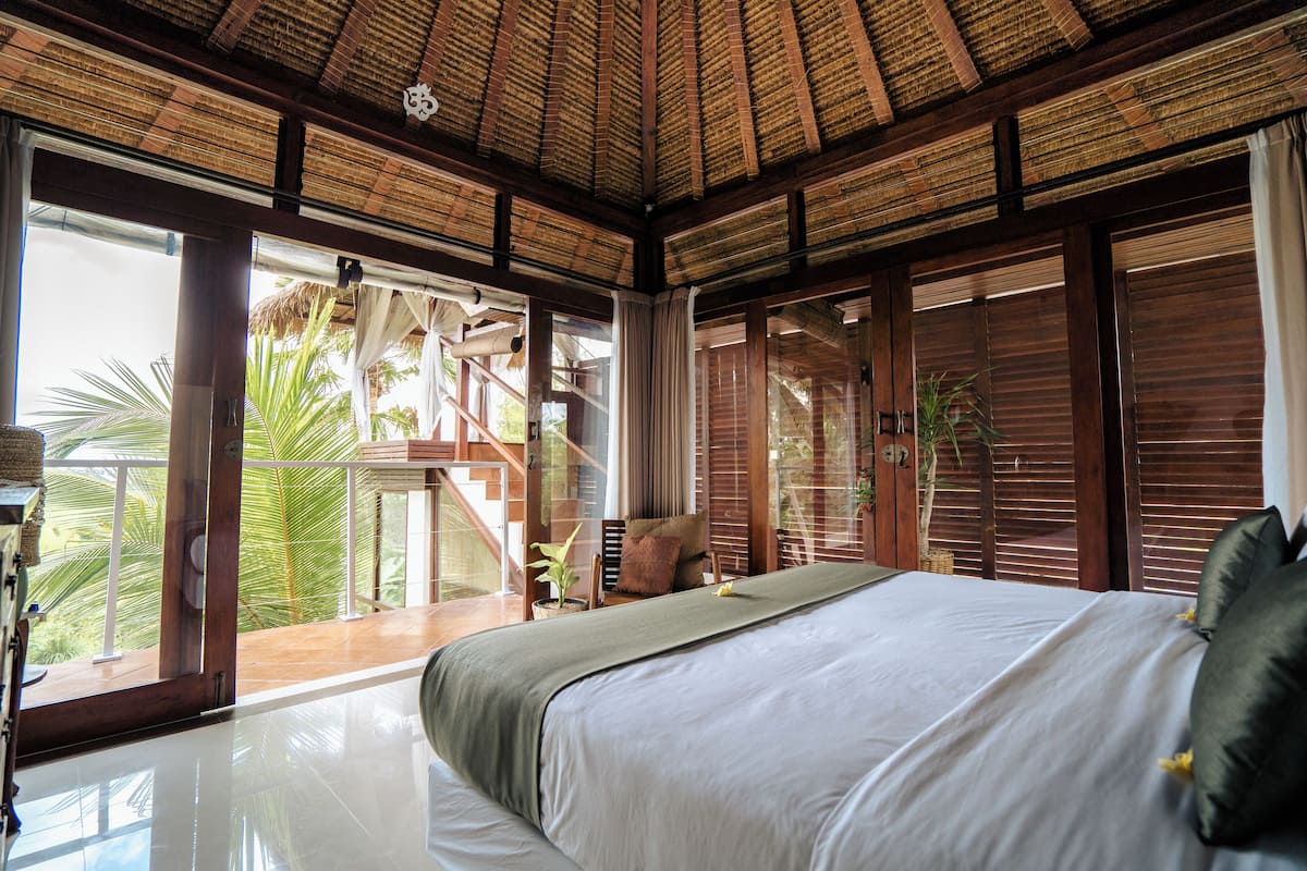 Quarto espaçoso com uma cama grande, teto de madeira e janelas amplas com vista para palmeiras.