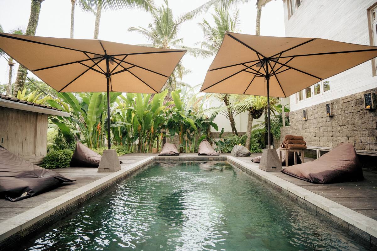 Uma área de piscina com dois guarda-sóis grandes, um espaço de estar com almofadas e uma parede de pedra.