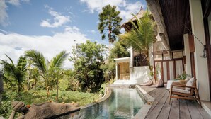 A pool with a stone sculpture, wooden deck, and a building with a thatched roof. - Basundari Retreat Bali (Ubud)