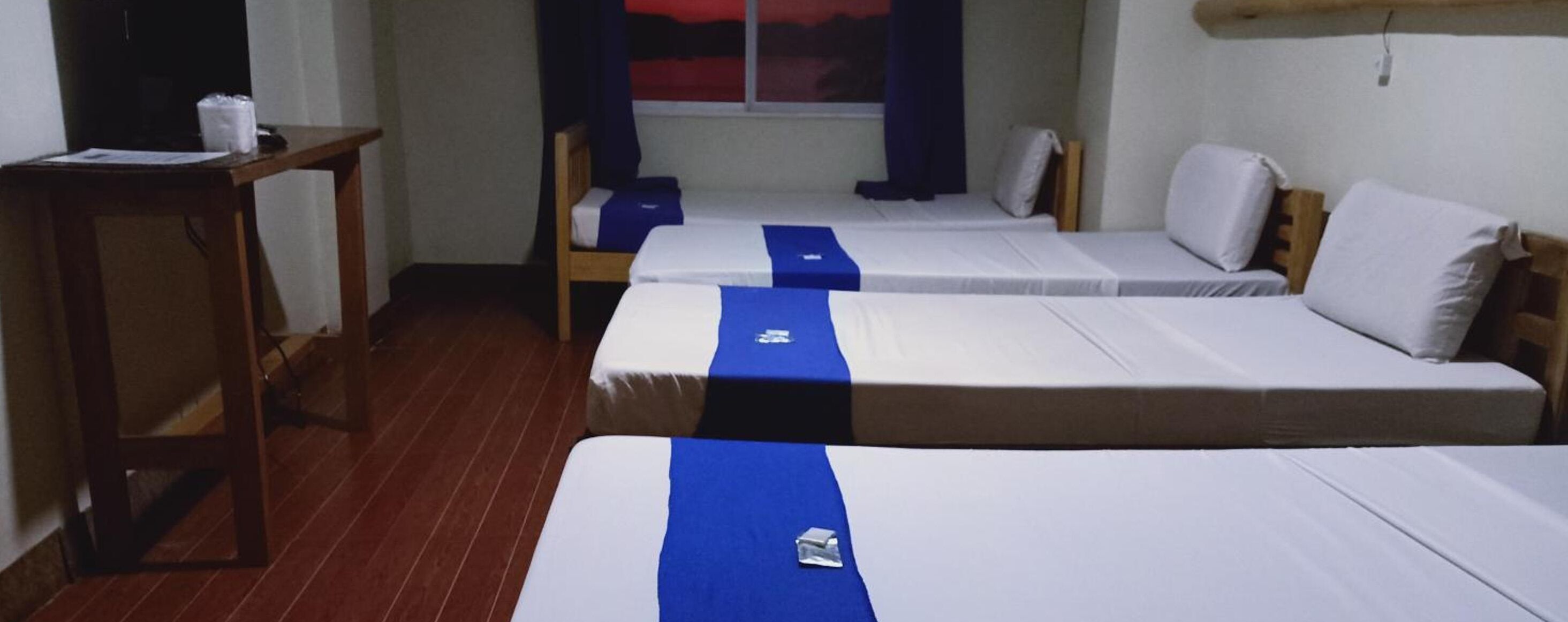 Quarto com três camas de solteiro, uma escrivaninha de madeira e janela com vista para o pôr do sol.