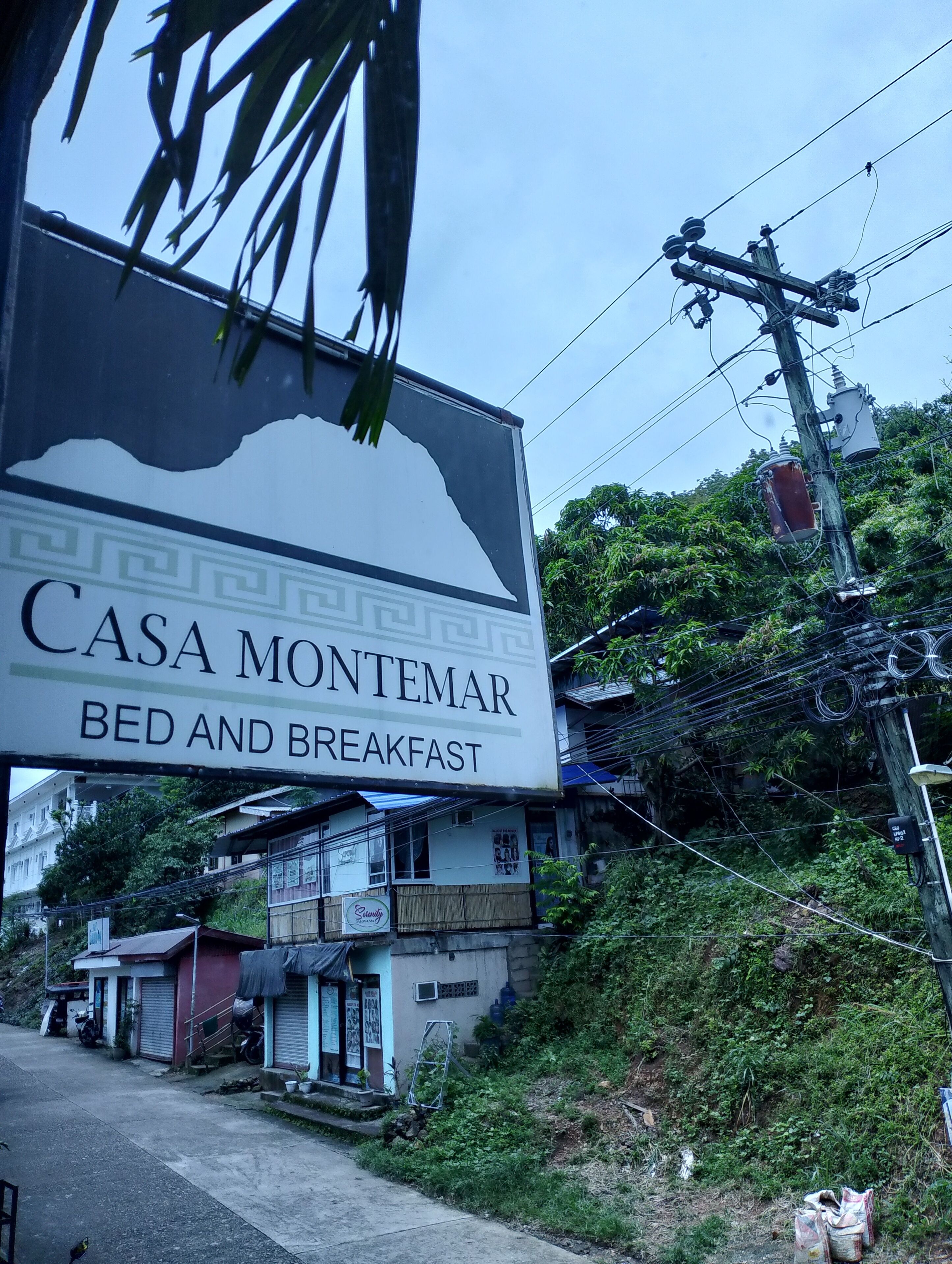 Schild für 'Casa Montemar Bed and Breakfast' mit Bergsilhouette, umgeben von Gebäuden und Vegetation.