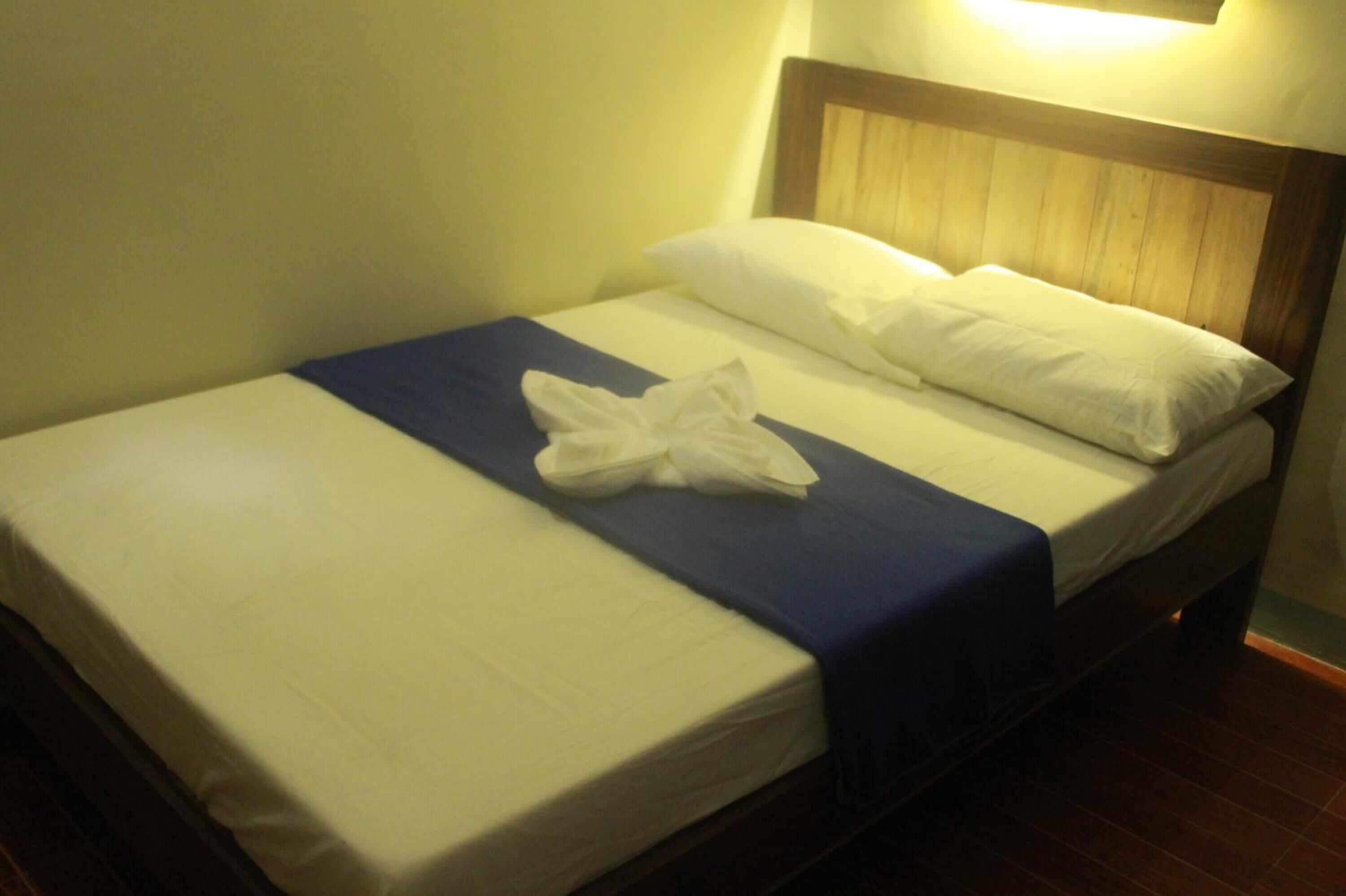 A neatly made bed with a blue bedspread and white linens, a wooden headboard, and a wall-mounted light fixture.