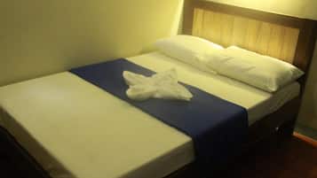 A neatly made bed with a blue bedspread and white linens, a wooden headboard, and a wall-mounted light fixture.