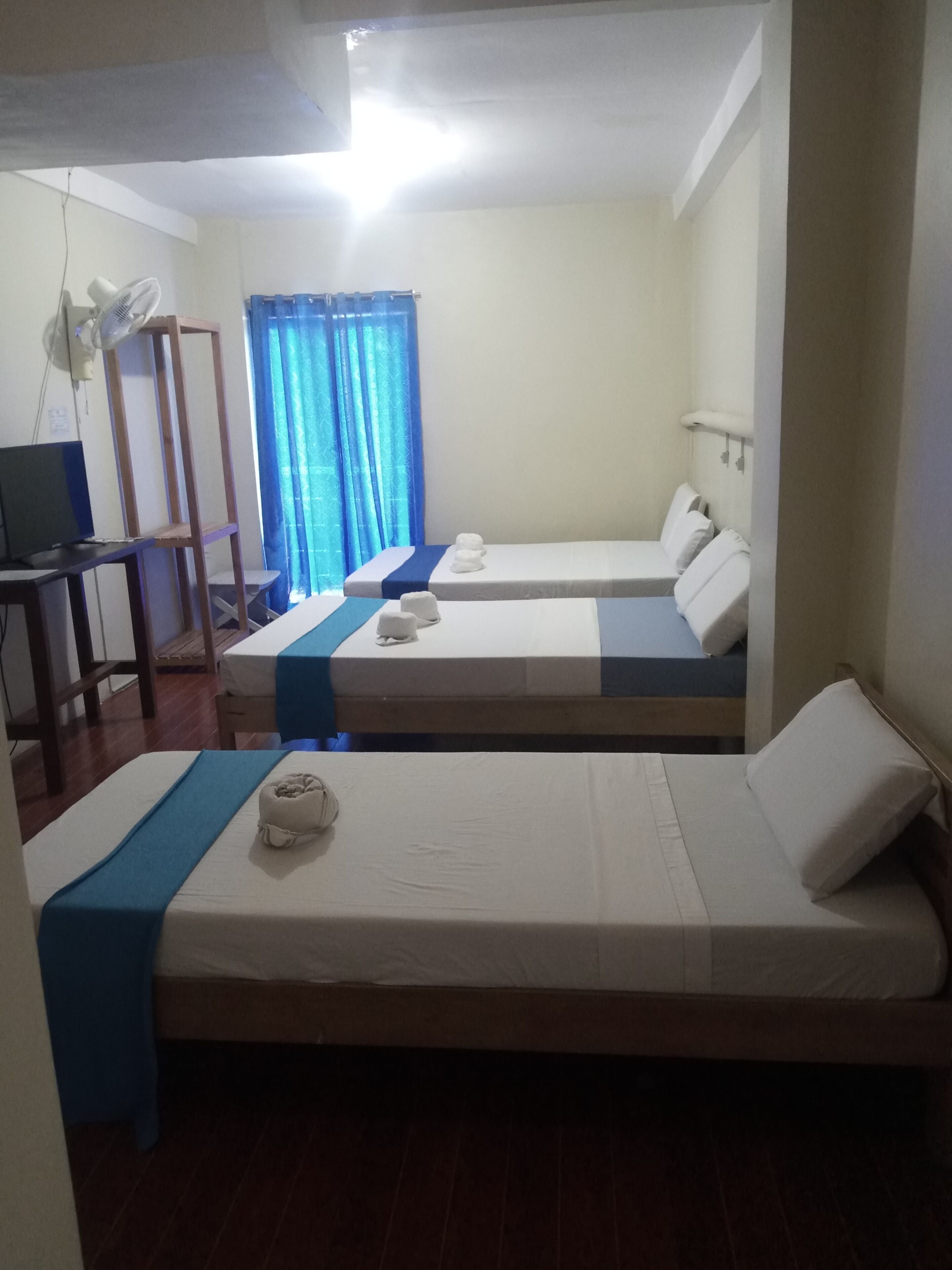 Habitación de hotel con dos camas individuales, un escritorio con una computadora, un ventilador y una ventana con cortinas.