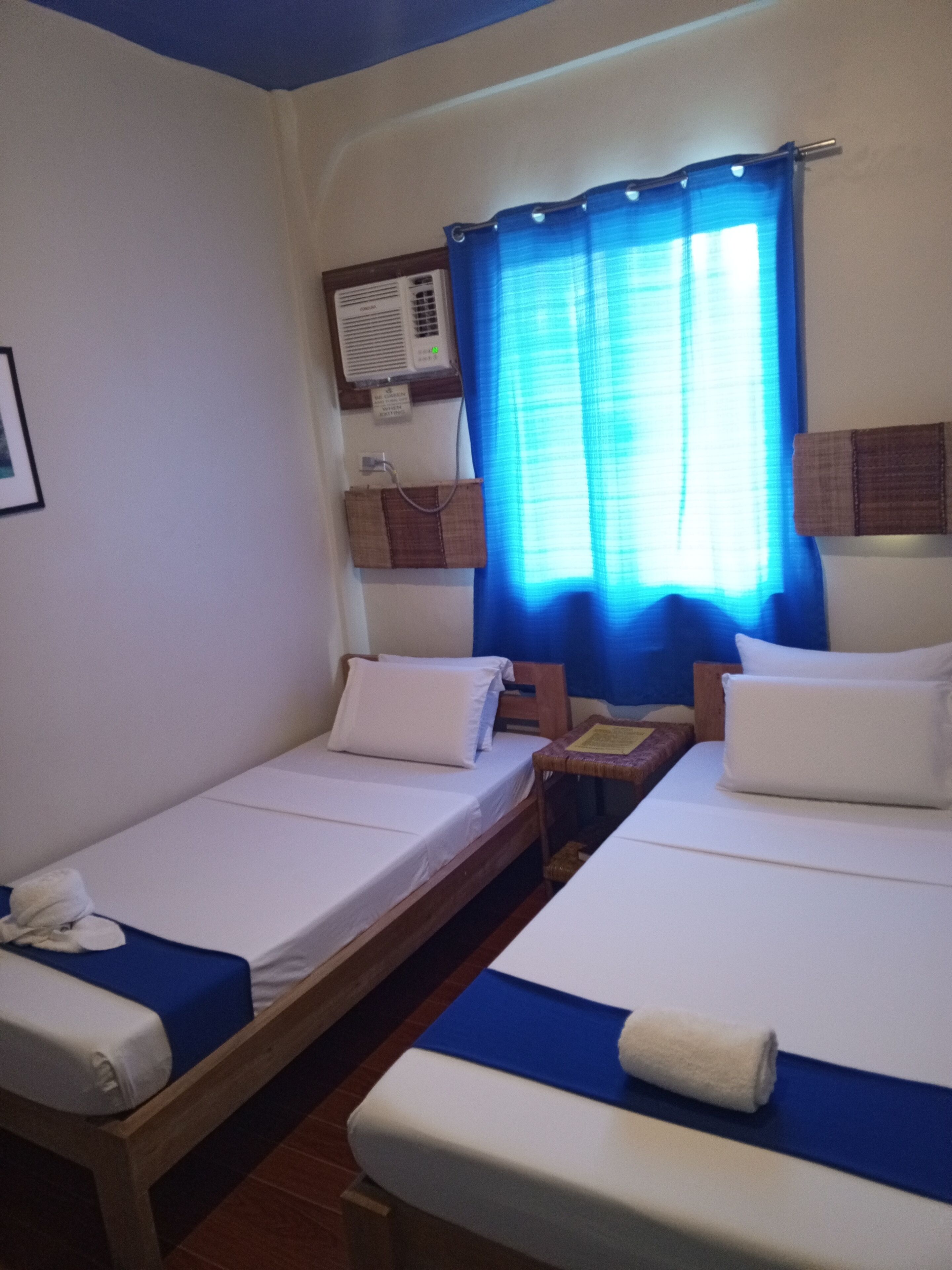 Una habitación con dos camas individuales, una mesita, aire acondicionado de pared y una ventana con cortinas azules.