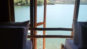 A room with a view of a lake and hills. There are two pillows on a bed.
