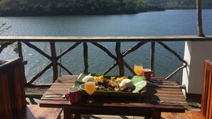 A wooden table with a meal and drinks on a deck overlooking a lake with hills in the background.