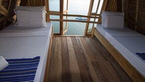 A room with two beds, wooden flooring, and a view of a body of water through a window.