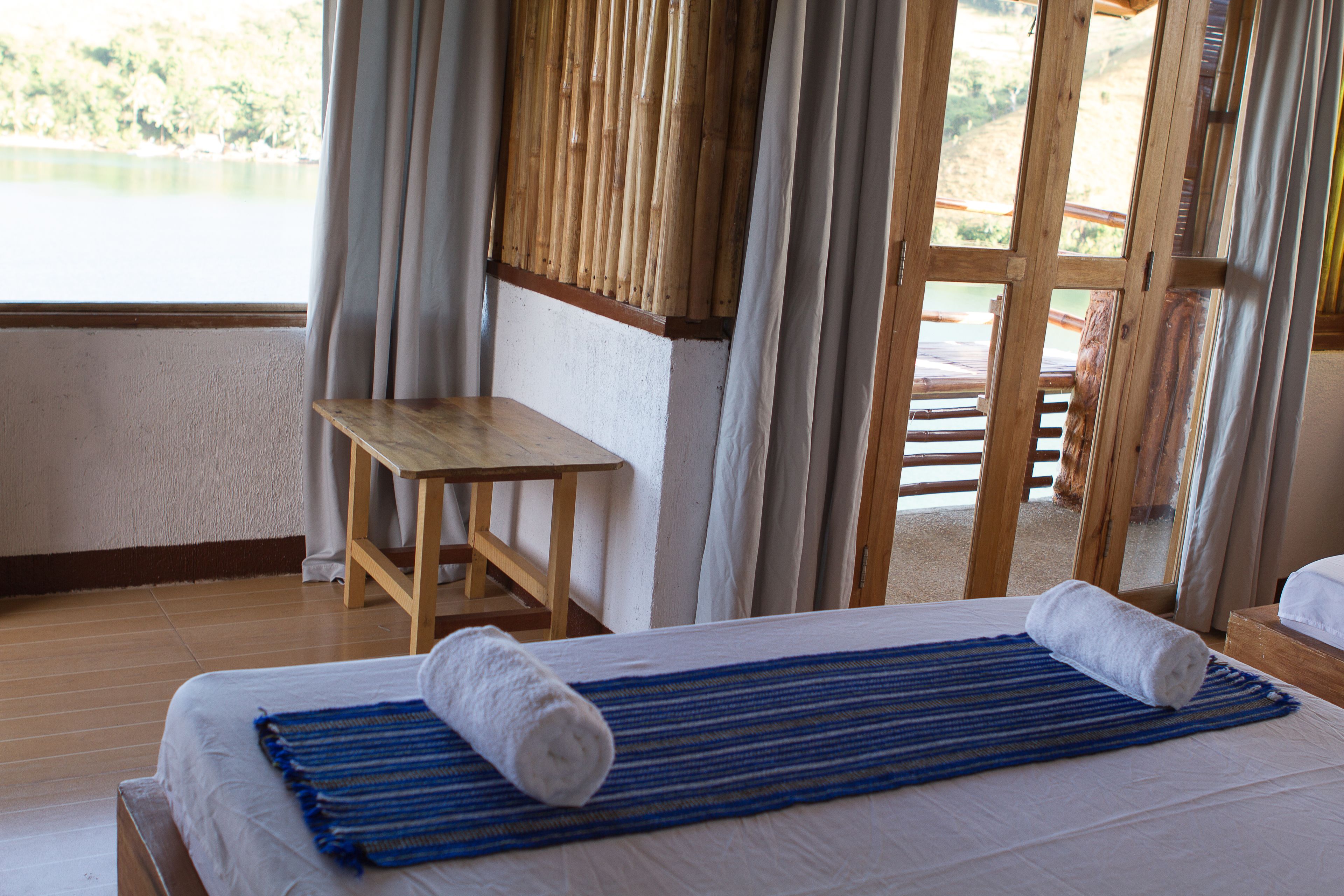 A hotel room with a bed, a wooden table, and a view of a lake through a large window.