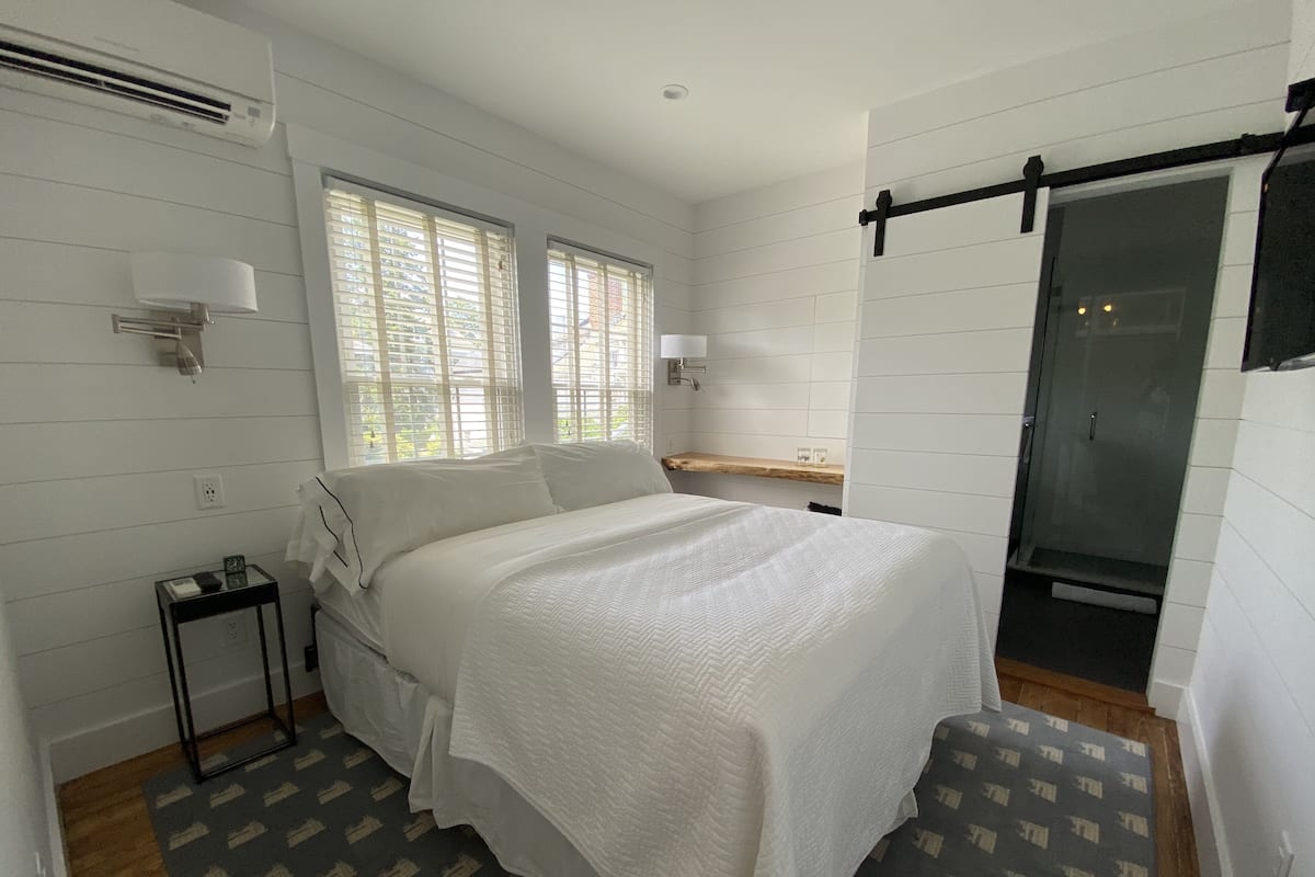 A bedroom with a bed, bedside tables, a nightstand, a window, a door, and a shower area.