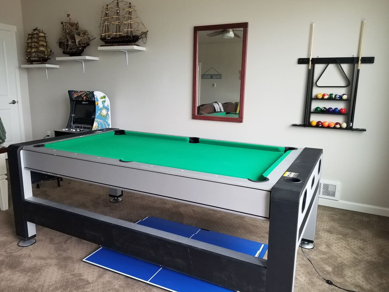 Ett biljardbord med en grön filt på bordsskivan, en blå filt på undersidan och en svart ram. Ovanför bordet finns tre hyllor där modellfartyg står utställda.