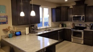 A modern kitchen with dark wood cabinets, stainless steel appliances, a large island, pendant lighting, and a view of the ocean through a window. - Beach Escape - Waldport (Waldport)