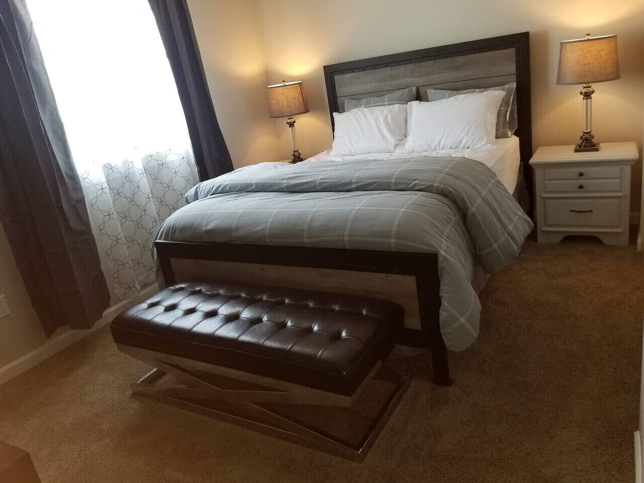 Une chambre à coucher avec un lit, deux tables de chevet avec des lampes, un banc et une fenêtre avec des rideaux.