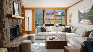 A cozy living room with a stone fireplace, a large window with a view of a mountain landscape, a white sofa, and a coffee table. - Villas at Snowmass Club - 1423 ~ Snowmass Club Access ~ (Snowmass Village)