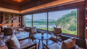 Habitación amplia con un gran ventanal que ofrece vistas de un río y montañas. Hay varias sillas y una mesita con un jarrón.