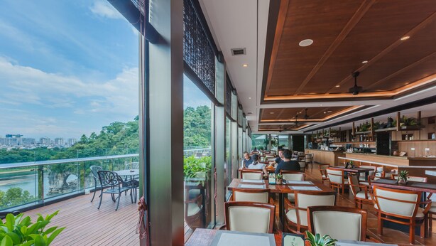 Restaurante con terraza al aire libre con vistas a un río, con mesas y sillas de madera, zona de bar y grandes ventanales que ofrecen vistas a la ciudad y al follaje.