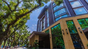 Un edificio moderno con fachada de vidrio, un acceso cubierto y un área ajardinada con árboles.