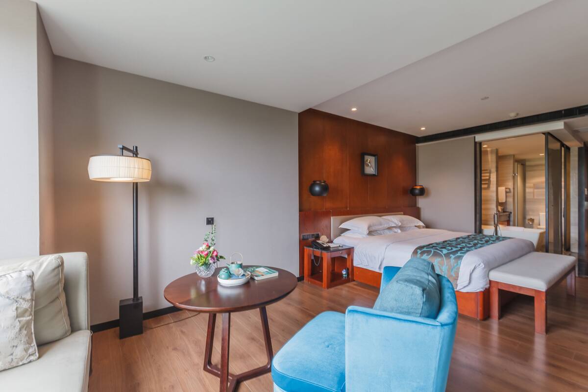 A modern hotel room with a bed, a sofa, a round table with a lamp, and a bedside table with a vase and teapot.
