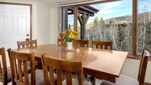 A dining area with a wooden table and chairs, a vase with flowers, and a view of trees and mountains through large windows. - Terracehouse 54 - Ski by Day, Soak by Night | Steps to Slopes & Hot Tub (Snowmass Village)