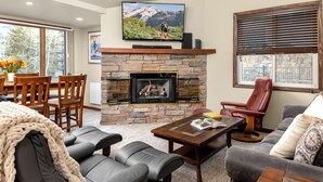 A living room with a stone fireplace, leather sofas, a dining area with wooden chairs, and a television mounted above the fireplace. - Terracehouse 54 - Ski by Day, Soak by Night | Steps to Slopes & Hot Tub (Snowmass Village)