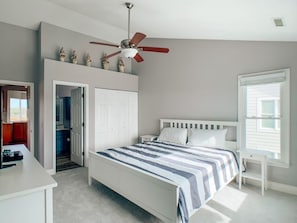 Une chambre à coucher comprenant un lit, un ventilateur de plafond, une fenêtre et une porte donnant sur une salle de bain.