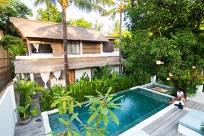 Un área junto a la piscina con una casa de techo de paja, abundante vegetación y una terraza de madera.