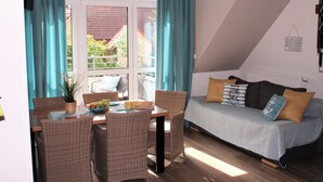 A dining area with wicker chairs and a table, a sofa with cushions, and a view through a window with curtains.