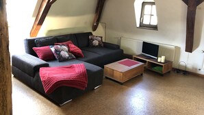 Un salon chaleureux de style loft, avec un canapé gris foncé, des coussins rouges et une table basse en bois. Un téléviseur à écran plat est fixé au mur, et un radiateur est visible.