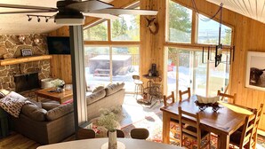 A cozy cabin interior with a stone fireplace, wooden walls, a dining area, and large windows offering a view of the outdoors. - WinterPark Chalet-3br/2ba. 2decks/hottub/mtn views. Skiing/Hiking/Biking nearby. (Tabernash)