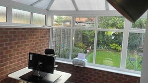 A conservatory with a desk and computer, large windows, and a view of a garden. - Charming Village house in Bramhall near Manchester (Bramhall)