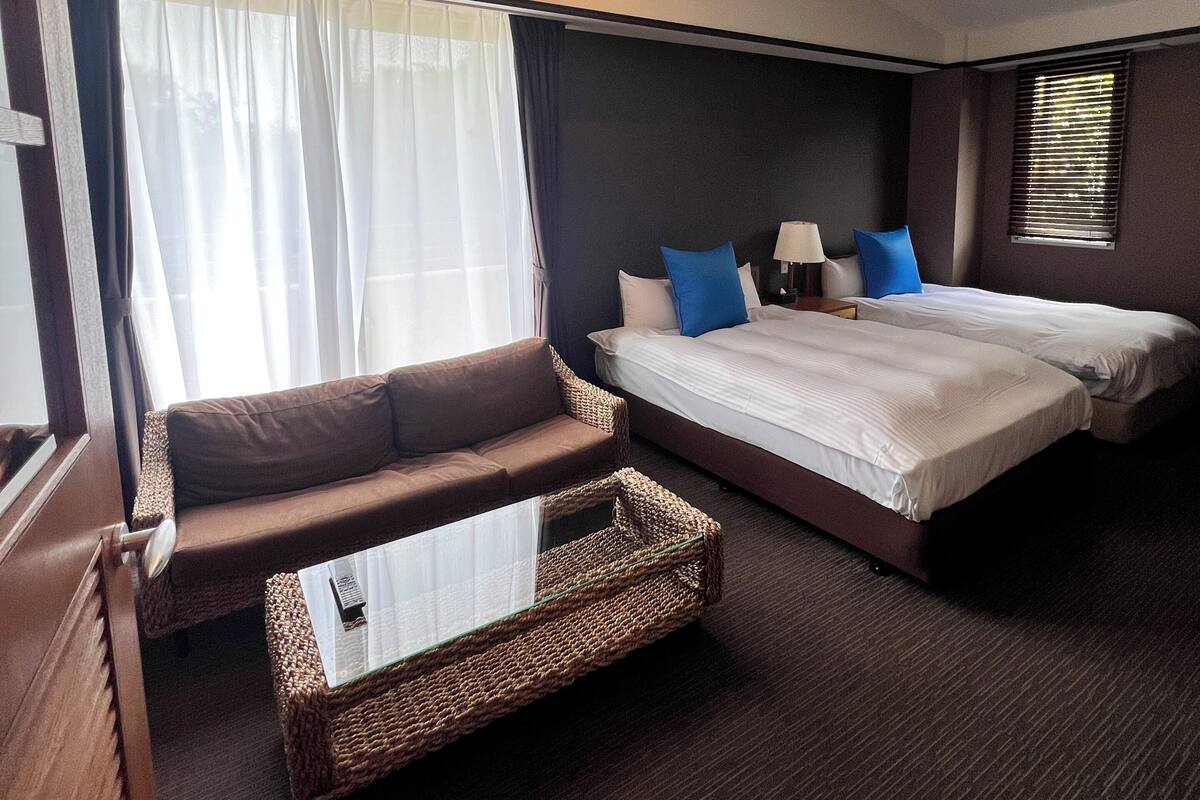Quarto de hotel com sofá, cama com duas almofadas, uma mesinha e janela com persianas.
