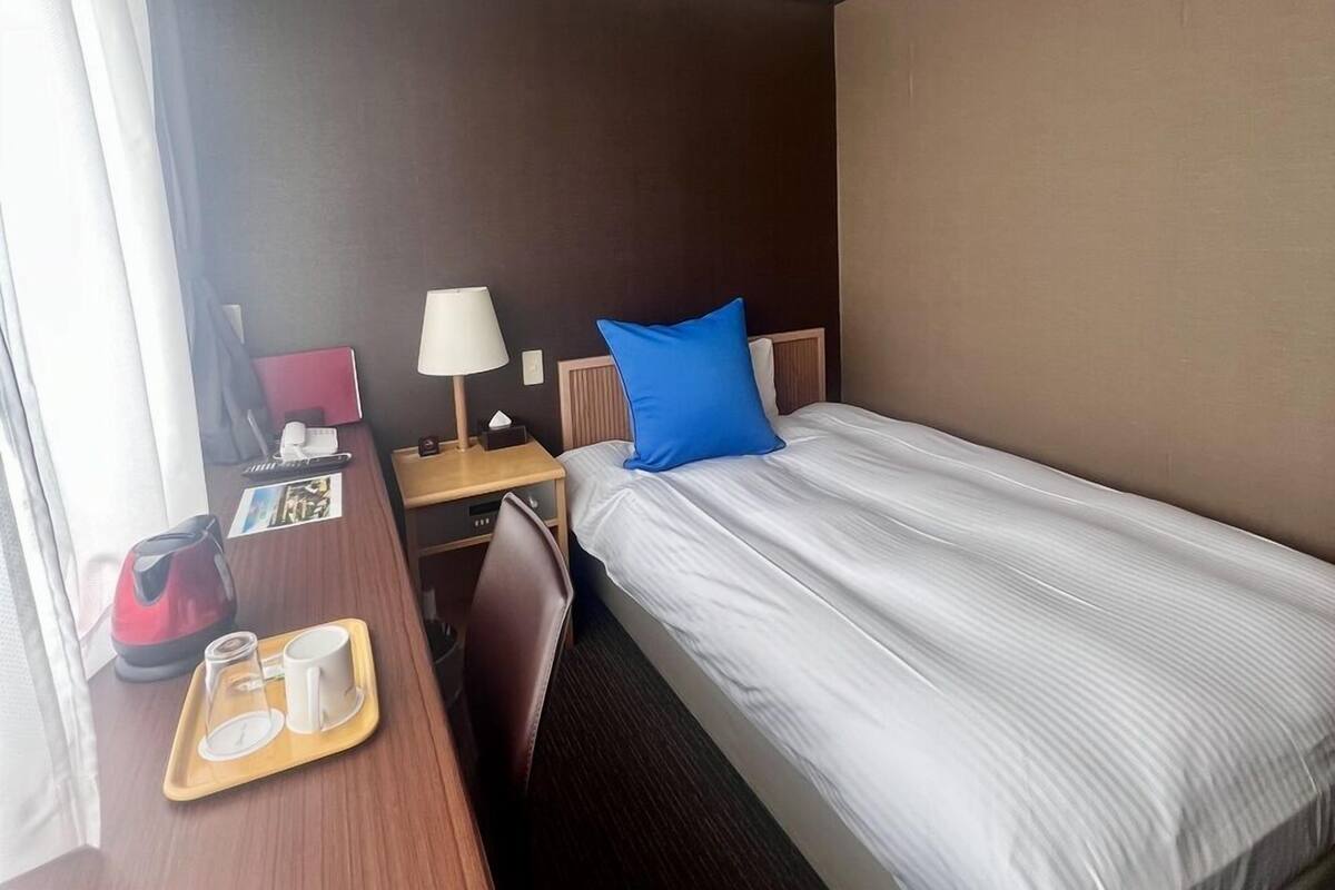 Quarto de hotel com cama, escrivaninha com luminária, cadeira, bandeja com xícara e chaleira, e janela com cortinas.