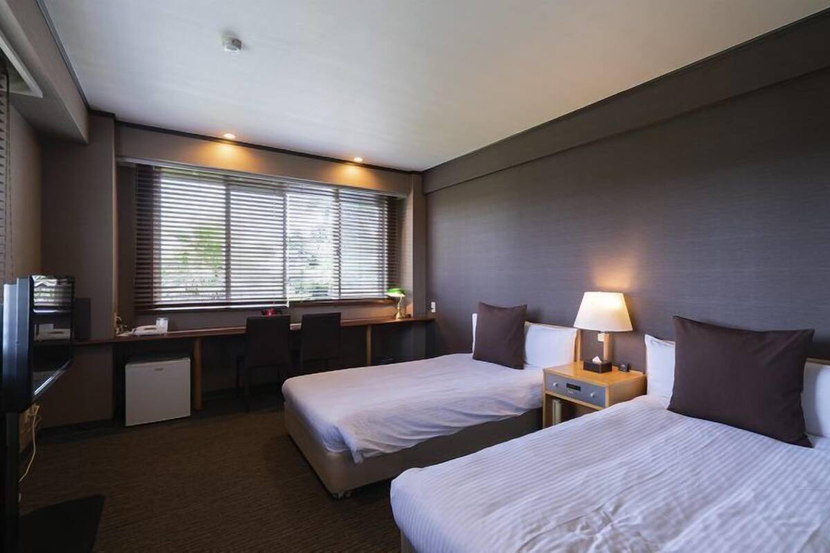 Quarto de hotel com duas camas, uma escrivaninha, uma cadeira, um pequeno frigorífico e uma janela com persianas.