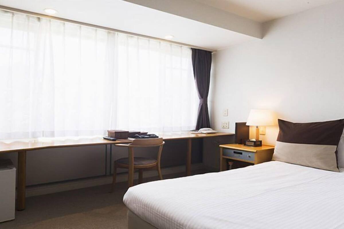 Quarto de hotel com cama, escrivaninha, cadeira, luminária e janela com cortinas.