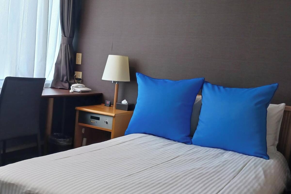 Quarto de hotel com cama, escrivaninha, luminária, cadeira e mesa de cabeceira.