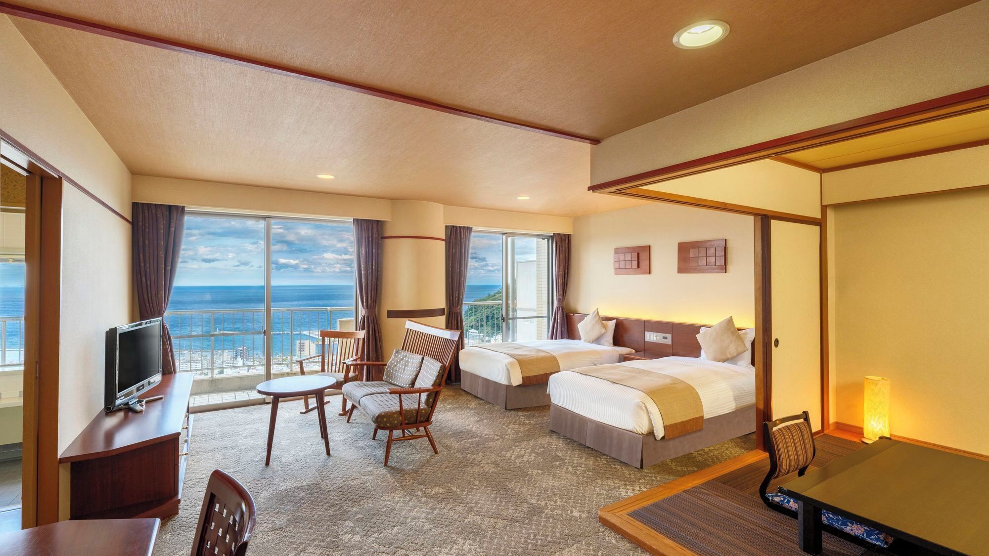 A hotel room with two beds, a desk with a TV, a chair, and a view of the ocean.