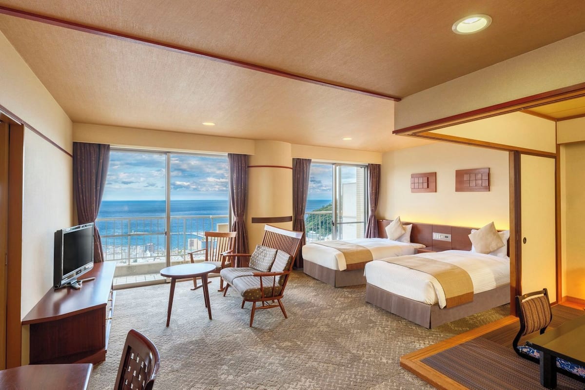 A hotel room with two beds, a desk with a TV, a chair, and a view of the ocean.