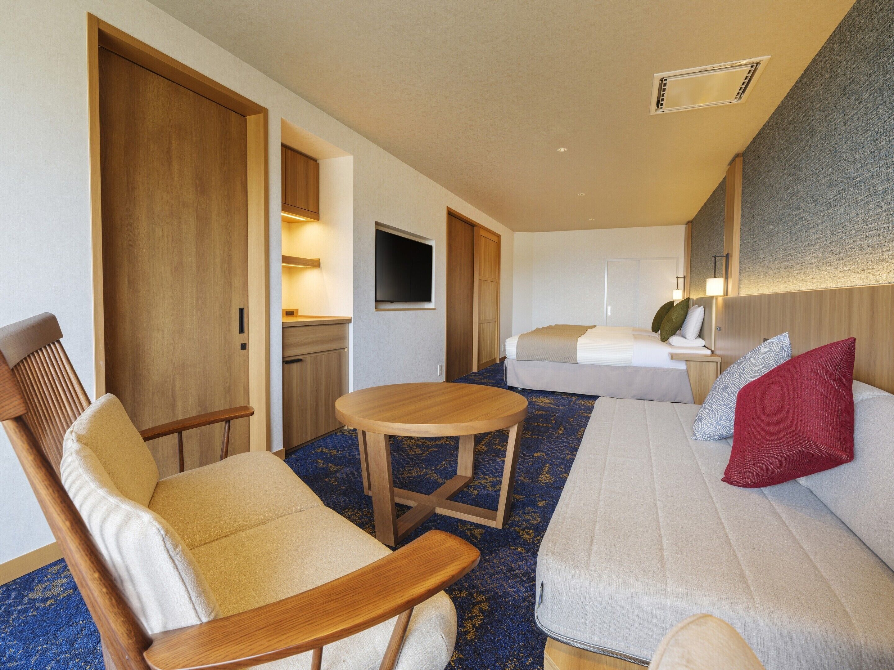 A hotel room with a sofa, a chair, a small table, a bed, a TV, and a bathroom with a shower and toilet.